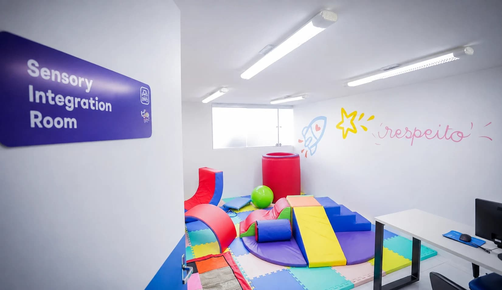 A sensory integration room: soft play shapes, a crash mat, floor tiles and a low ramp, under even lighting.